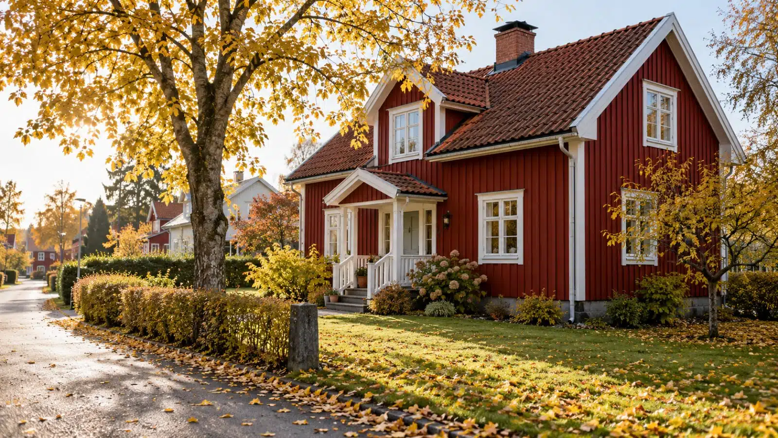 En röd trävilla med vita knutar och en trädgård med gula höstlöv längs en lugn villagata.